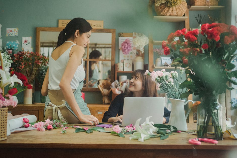 A floral workshop scene featuring two women in a brightly lit flower shop. The woman on the left, standing, has dark hair tied back and is wearing a sleeveless white top with a beige apron tied around