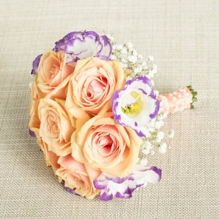 This exquisite bouquet, reminiscent of Golders Green's tranquil gardens, invites one to experience its every detail: satiny peach and apricot roses, their petals soft as the skin of ripe fruit from the local farmers' market, massed together in lush abundance. Each rose reveals a heart of subtle pink and is surrounded by creamy exteriors, offering a sense of tenderness and freshness. Artful interludes of white lisianthus, each bloom rimmed with a dramatic swipe of purple, lend a flourish of contrast, mirroring the creative vibrancy found in Golders Green's bustling community. Threaded throughout, baby's breath forms a light, airy weave that gives the bouquet a cloud-like, almost ethereal quality-perfect for elevating the spirit of any celebration. The compact, rounded shape echoes classic wedding traditions, while a peachy-pink satin ribbon, studded with elegant pearl beads, envelops the stems, providing a tactile flourish and secure grip. This bouquet captures the essence of occasion, whether chosen for a Golders Green wedding, engagement, or cherished garden party near Hampstead Garden Suburb, reflecting a harmonious blend of romance, sophistication, and modern sensibility in every detail.