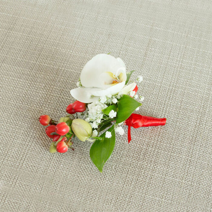 An elegant floral boutonniere is captured from above, lying on a subtly textured, light beige woven fabric that looks like natural linen or fine burlap, its neutral hue providing a soft, homely backdrop reminiscent of a carefully set Golders Green reception table. At its centre sits a pristine white Phalaenopsis orchid, the petals broad, smooth, and slightly curved, as though gently cupping the air. The orchid's throat glows with a delicate wash of creamy yellow and soft orange, a small, warm contrast that draws the eye inward. Clustering closely around this central bloom are tiny white flowers of baby's breath, their airy, cloud-like sprays filling the spaces between petals and adding a sense of lightness and romance, like a mist of blossoms surrounding the orchid. On one side of the arrangement, a group of glossy, round red berries adds a cheerful burst of colour, their surfaces almost reflective, suggesting freshness and a hint of festivity often associated with winter weddings or Christmas parties in North West London. Nestled among them are a few paler green, berry-like buds, a gentle nod to the cycle of growth. Interwoven with the flowers and berries are rich green leaves in different forms - some broad and pointed, others slender and refined - which frame the central orchid and anchor the composition with natural structure. The stems are carefully bound at the base with a vivid red satin ribbon, tightly wrapped and finished in a neat, compact knot with short tails, the fabric catching the light with a soft sheen. The boutonniere feels poised and ready to be pinned to a lapel, perhaps for a groom stepping out near Golders Green Station or a guest arriving at a formal event along Temple Fortune, conveying a mood of polished celebration and thoughtful craftsmanship.