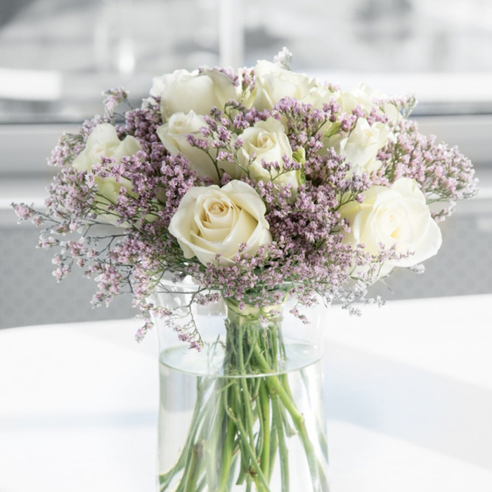 A refined floral arrangement fills the frame, showcasing a dozen or so creamy white roses nestled among clouds of pale lavender-pink filler flowers, all gathered in a sleek glass vase that would not look out of place in a tranquil Golders Green sitting room. The roses are captured at their peak, their petals fully opened into large, rounded blooms that reveal intricate spiral centres and slightly ruffled edges, like soft folds of silk. Their colour is not a stark white, but a gentle cream that deepens to a faint ivory at the core, lending a sense of warmth and intimacy to the composition. Surrounding these central blossoms are plentiful stems of tiny, clustered flowers-likely limonium or statice-in muted shades of lilac, lavender, and dusky pink. These smaller florets form a delicate haze around the roses, their fine, papery texture providing subtle contrast and creating an almost cloud-like frame. The bouquet is anchored in a tall, cylindrical vase of completely clear glass, allowing a clean view of the straight, green stems gathered together and submerged in transparent water. The water line sits neatly below the blooms, reinforcing the feeling of freshness and careful preparation. The vase rests on a smooth, white surface that gently reflects the light, while the background is softly blurred into pale tones, reminiscent of a quiet interior near Golders Green station, where daylight drifts through sheer curtains. The lighting is soft and even, with no harsh shadows, enhancing the calm, contemplative mood. This image conveys serenity, dignity, and understated luxury, making it an apt visual representation of a sympathy bouquet designed to bring comfort to bereaved families and friends across Golders Green and the nearby Finchley Road area.