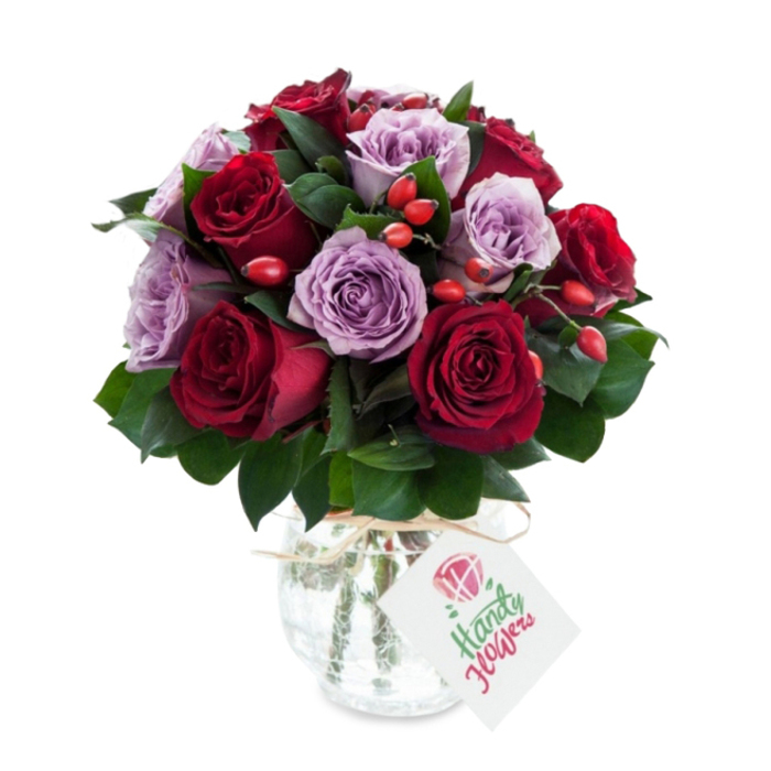 This compact, abundant bouquet juxtaposes rich, velvety red roses in tones of crimson and burgundy with softer lavender and lilac roses, arranged in a dense, rounded silhouette so the eye moves smoothly from dark to pale and back again. The roses are at peak bloom, their layered petals open and slightly rolled at the edges, suggesting freshness and careful timing at the florist's table. Interspersed foliage of deep green - suggesting Ruscus or Salal - provides structural contrast and a cool, matte foil for the petal sheen, while small bright orange-red rose hips or berry clusters are dotted through the heads to add tactile contrast and a natural, slightly rustic accent. The stems rest in a decorative mercury-style glass vase: reflective, antiqued and subtly mottled, rounded at the base and narrowing at the neck, finished with a natural raffia or twine tie that softens the metallic look. A neat white rectangular gift tag hangs from the neck, carrying a tiny floral sketch and space for a handwritten note, making the piece ready for gifting. Photographed on a stark white background, the arrangement's colours and textures are read clearly - an elegant choice for an anniversary table, a thoughtful birthday surprise, or a special delivery to a Golders Green flat after a walk from the Tube station, showcasing the kind of considered composition local florists rely on when selecting each bloom.