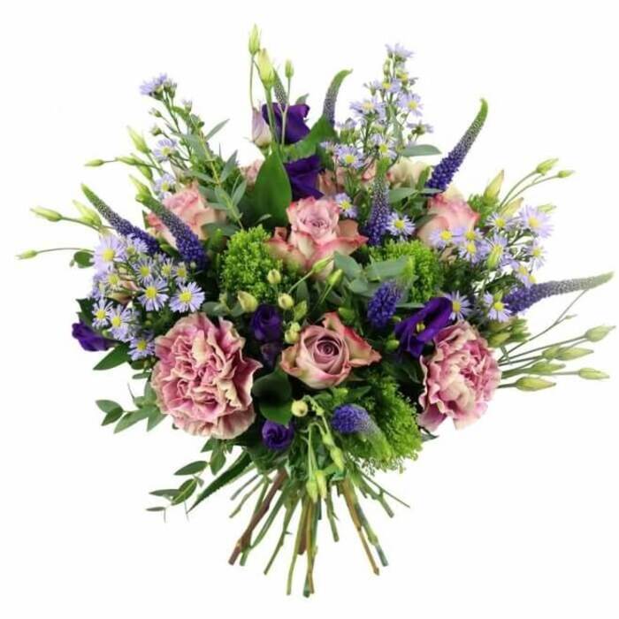 Captured from a slightly overhead angle against a bright white background, this hand-tied bouquet feels like a carefully composed painting brought to life in a Golders Green flower shop. The centre of the design is filled with soft pink and mauve roses, each at a different stage of bloom, from tight buds hinting at future beauty to open flowers that reveal intricate spirals of petals. Their colouring shifts subtly from deeper rosy hearts to pale, almost ivory outer edges, lending a romantic, vintage quality. Nestled between them, plush carnations in tones of dusty rose and purple-mauve add a frilled, voluminous texture, ensuring the bouquet looks full and luxurious from every angle. Rising with poise among the rounder blooms, slender spikes of deep lavender-blue Veronica create elegant vertical accents, adding structure and a contemporary feel that suits modern homes around Golders Green Road and nearby Hampstead Garden Suburb. Smaller, starry aster-like flowers in light lilac and periwinkle, each with a muted yellow centre, form a soft, cloud-like filler, while richer purple lisianthus blooms and buds offer pockets of depth and mystery. Lush greenery weaves throughout: silvery eucalyptus leaves, brighter lime-green filler, and other foliage combine to frame the flowers and intensify their colours, much like fresh foliage found in local parks. The stems at the base are cleanly trimmed and gathered into a tight, hand-tied bundle, indicating the bouquet is ready to slip into a vase after delivery to a home near Brent Cross or a cosy café off Finchley Road. The overall mood is one of refined elegance and heartfelt sentiment, ideal for marking milestones, sending congratulations, or bringing a touch of luxury to a Golders Green living room.