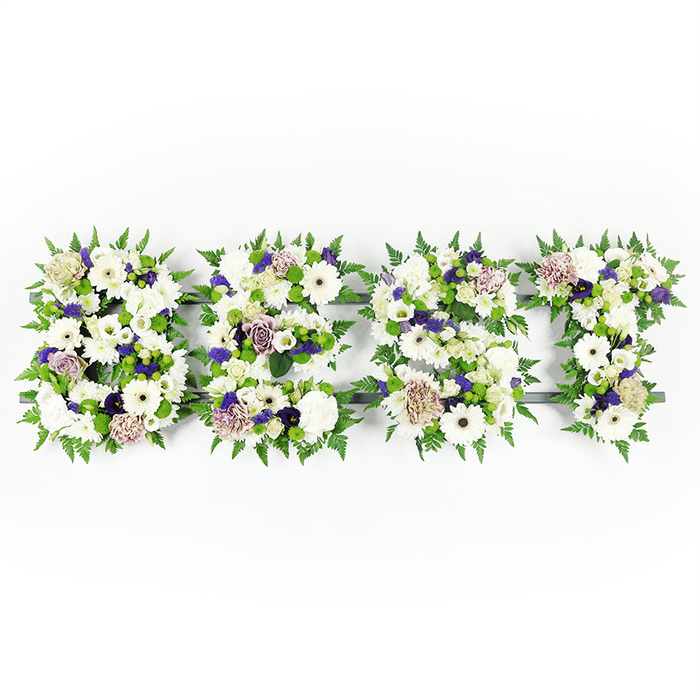 A horizontal, hand-crafted floral letter tribute spelling BEST, presented against a pristine white backdrop where every petal and frond is visible in gentle, natural light. Each uppercase letter-B, E, S and T-is densely composed of luminous white gerbera daisies with their dark centres, full white carnations, and delicate white lisianthus, the varied whites working together like pages in a softly bound book of memories. Soft mauve and lavender roses are threaded through the letters, their velvety tones adding a measured warmth that balances the purity of the whites, while smaller clusters of deeper purple filler lend a quiet depth and reverence. Bright green button chrysanthemums and compact green buds sit within the floral layers, offering pops of verdant cheer and a pleasing sculptural texture. Edge-defining fern fronds create a feathery border around each letter, their fragile curves bringing a natural rhythm that makes the composition breathe. The dark grey structural base beneath the blooms is discreetly visible in places, the contrast lending precision to the letter shapes without taking from the soft floral impression. This is a sympathy-style tribute that reads as both an artistic statement and a personal message, appropriate for memorial services or graveside remembrances around Golders Green, from the lanes by Hoop Lane to the walkways near Hampstead Heath. It carries the simple, composed dignity of a floral farewell crafted to honour a life with love and restraint.
