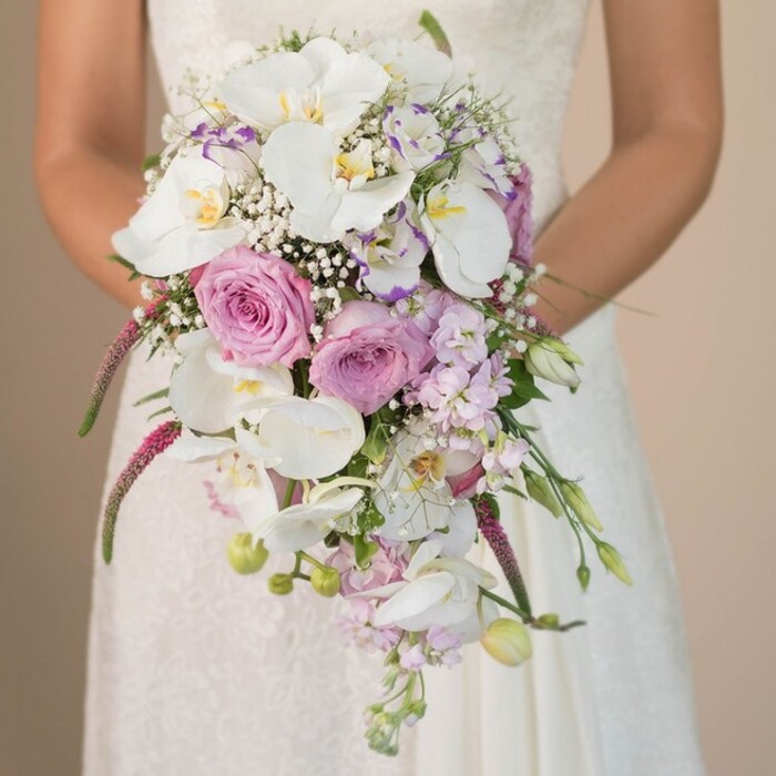 Nestled within the refined calm of Golders Green, a close-up image unfolds a bride's gentle posture as she holds a spectacular cascading bouquet that truly embodies luxury and romance. Her white gown, touched with delicate lacework, forms a gentle contrast to the lush waterfall of florals spilling from her embrace. At the heart of the bouquet, broad, pristine Phalaenopsis orchids with subtle golden centers instantly suggest a sense of opulence; their elegant forms catch the soft light that drifts across the scene. Clusters of pink and lavender roses, many at the blush of full bloom, lend a timeless softness, their velvety petals inviting the gaze and imagination alike. Ethereal gypsophila-baby's breath-appears as fine, airy wisps, weaving between blooms to create a dreamy, cloudlike effect. Slender spikes of magenta Amaranthus cascade delicately, adding a dramatic sweep that mimics the natural fall of flowers over Hampstead Heath in spring. Flecks of green foliage and curled unopened buds peek out, hinting at freshness and thoughtful composition by an expert Golders Green florist. The muted background-a nod to the understated elegance of local architecture-ensures nothing distracts from the bouquet's intricate splendour. Every element, from the draping orchids to the dusting of lavender and blush, infuses the image with a spirit of celebration and the tender hopefulness of marriage. This arrangement is more than flowers: it's a crafted ode to enduring love, perfect for a wedding set in the leafy, welcoming streets of NW11.