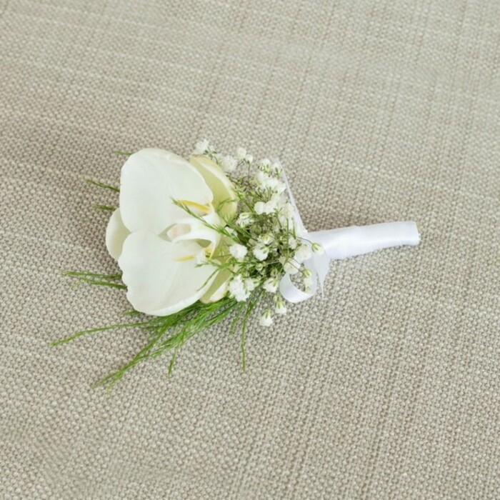 A meticulously crafted white orchid boutonniere is displayed on a light beige, woven fabric that looks like finely textured linen, its neutral tone allowing every floral detail to shine. The main bloom, a pristine Phalaenopsis orchid, opens in a graceful arc, its broad, slightly glossy petals radiating pure white while a tender wash of pale yellow glows from the centre of the flower. This gentle contrast adds warmth and depth, evoking the soft, golden light you might notice over nearby Golders Hill Park in the late afternoon. Surrounding the orchid, clusters of delicate baby's breath form a delicate halo, hundreds of tiny white blossoms gathering into airy clouds that soften the silhouette of the piece. Nestled around and beneath these miniature blooms are slender strands of bright green foliage, their fine, lacy shape bringing a whisper of woodland texture, as if borrowed from a shaded garden off North End Road. The stems are cleanly aligned and tightly bound with a wrap of smooth white satin ribbon, which clasps the arrangement in a crisp, polished band. Near the top, the florist has tied a small, neat bow, giving the boutonniere a refined finishing touch that speaks to practiced hands and an eye for balance. The lighting in the image is soft and even, with no harsh shadows, enhancing the tranquil mood and allowing the subtle sheen of the orchid and ribbon to emerge gently. The boutonniere feels perfectly suited to a groom or groomsman on a Golders Green wedding day, perhaps stepping out from a family home onto a quiet residential street. Its overall impression is one of classic elegance, understated luxury, and the promise of new beginnings, ideal for formal celebrations where calm beauty and thoughtful design are cherished. Crafted in the spirit of local artistry found at Flowers Golders Green, it embodies the neighbourhood's blend of tradition and modern simplicity.