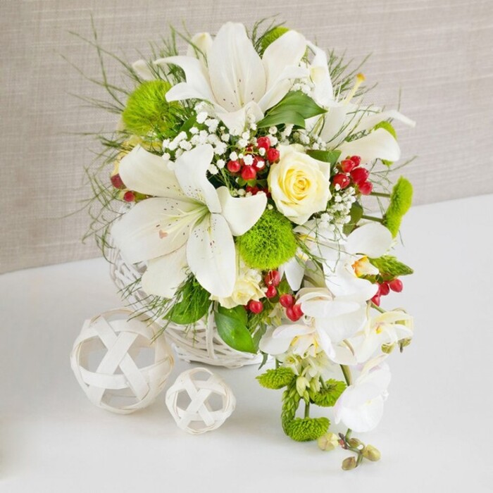 Displayed on a pristine white surface with a softly textured beige backdrop, this lavish floral arrangement captures the quiet sophistication of a Golders Green celebration. The flowers are artfully gathered into a low, round white wicker basket, its tightly woven strands giving a cosy, handcrafted character that wouldn't feel out of place in a stylish home near North End Road or along the leafy streets by Golders Hill Park. Rising from the center, several grand white Oriental lilies open fully, their velvety petals arching outward and revealing fine freckles and prominent stamens dusted with warm pollen. Just above them, a single buttery cream rose nestles confidently, its tightly layered petals forming a neat rosette and drawing the eye like a gentle focal point. From the right side, a line of elegant white phalaenopsis orchids spills downward in a soft cascade, each bloom flat and wide, with a sculpted, almost butterfly-like shape that introduces a sense of fluid motion to the otherwise compact arrangement. Surrounding the main blooms, lush green foliage in varying shapes-feathery fronds, slim blades, and slightly broader leaves-creates depth and freshness, while bright green pom-pom dianthus add playful spheres of mossy texture. Tiny sprays of white gypsophila weave a delicate veil between the larger heads, and glossy ruby-red berries punctuate the composition, suggesting warmth, celebration, and a hint of seasonal festivity. The lighting is gentle and diffuse, enhancing the creamy whites and cool greens, and making the entire design feel calm and inviting. To the left, two white decorative wicker balls, one larger, one smaller, echo the basket's weave and complete the image as a polished, contemporary wedding or dinner party centerpiece ready for same-day delivery across Golders Green.