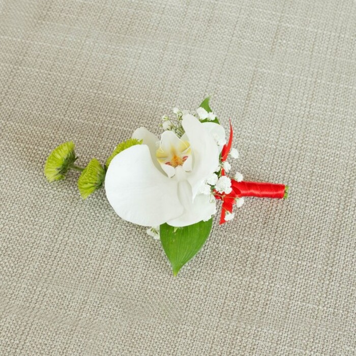 An artfully crafted boutonniere lies on a softly textured, light beige linen surface, the fabric's subtle, natural weave evoking a calm, understated backdrop like a reception table in a Golders Green venue before the guests arrive. At the centre is a pristine white phalaenopsis orchid bloom, its broad, gently curved petals forming a serene, symmetrical shape that draws the eye immediately. The orchid's throat reveals delicate details: a wash of soft yellow that deepens into refined reddish-orange or light brown markings, giving the flower a sense of depth and character, as if it has its own quiet story to tell. Nestled snugly around this central blossom are dainty clusters of white baby's breath, their tiny, cloud-like flowers adding a whisper of texture and an almost ethereal softness. To the left, two perfectly round lime-green chrysanthemum buds stand out, their intricate, slightly spiky surfaces offering a crisp, contemporary contrast to the orchid's smoothness, reminiscent of fresh greenery in the gardens off Finchley Road. Beneath the flowers, a broad, dark green leaf serves as a natural base, its rich tone grounding the design, while a smaller, deeper green leaf peeks out near the stems. The stems themselves are wrapped with meticulous precision in a vivid crimson red satin ribbon, pulled taut and smooth, culminating in a neat, modest knot that introduces a bold, joyful accent. The neutral, linen-like background ensures that every colour and texture-from the snowy white petals to the bright green buds and ribbon's glossy sheen-stands clearly defined. The overall impression is one of refined celebration, perfect for a groom, groomsman, or honoured guest at a North West London wedding, ready to be pinned to a suit as a subtle but meaningful statement piece.