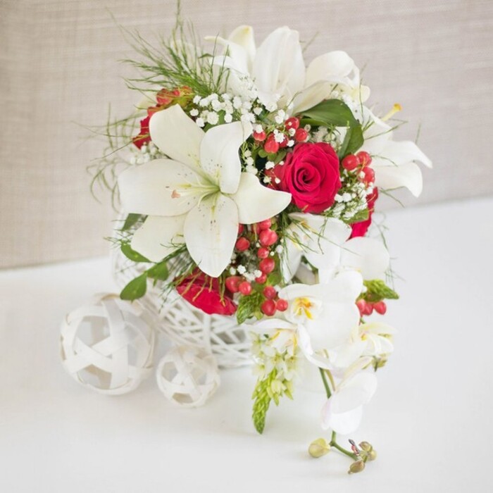 An elegant wedding-style table arrangement in white and red is carefully composed on a smooth white surface, as if prepared for a special celebration in a Golders Green home or boutique venue off Golders Green Road. The flowers are arranged in a distinctive white basket-style container with open, lattice-like weaving, giving it a light, airy profile that contrasts beautifully with the lush blooms it holds. Dominating the composition are several large white oriental lilies, their wide, starry blooms open to reveal prominent stamens tipped with warm brown pollen. These lilies feel both sculptural and soft, their petals slightly curved and catching the gentle light. Interwoven with them are velvety red roses, rich in colour and tightly petalled, bringing an unmistakable romantic energy to the piece. Fine sprays of baby's breath are sprinkled throughout, their tiny white blooms adding delicate texture that resembles a soft mist over the main flowers. Clusters of shiny red hypericum berries nestle among slender green foliage, the bright berries adding an uplifting, festive note that would be at home on a winter wedding table or seasonal dinner near Hampstead Heath. To the right, an elegant spray of white phalaenopsis orchids cascades gracefully downwards from the main arrangement, each bloom with a pale yellow and faint pink centre, enhancing the sense of flow and luxury. On the left of the bouquet, three decorative white woven spheres sit casually on the table, subtly mirroring the container's material and shape and completing the carefully curated tableau. The background is a neutral, softly textured beige fabric, which keeps the focus firmly on the sophisticated colour palette and composed beauty of the design.