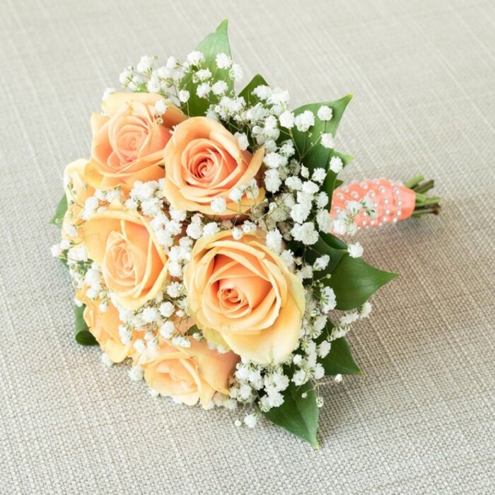 Reminiscent of a romantic celebration set in the heart of Golders Green, this hand-tied bridesmaid bouquet is a celebration of refined floral design and classic elegance. Gently resting across a subtly textured beige linen-perhaps echoing tablecloths found in stylish local venues-the bouquet showcases approximately seven perfectly bloomed roses in captivating shades of soft peach and pale apricot, their velvety petals gradually unfurling in delicate layers. Interlaced among the central roses are generous sprigs of pure white baby's breath, lending the composition an ethereal, almost cloudlike grace, beautifully balancing the warm vibrancy of the roses. Dark emerald leaves weave through the flowers, providing a touch of depth and a whisper of nature, an homage to the leafy streets and green spaces surrounding Golders Green. The stems are neatly encased in rich coral-peach satin ribbon, which elegantly spirals down to the base, adorned with a continuous row of luminous faux pearls. These pearls reflect the soft, diffused daylight typical of a Golders Green morning, adding a gentle shimmer and a sense of luxury. The entire bouquet is illuminated by flattering, even light, bringing its intricate textures and harmonious colour palette into full focus. Crafted with the artistry refined by years of serving local weddings and gatherings, this bouquet embodies timeless romance and is a treasured choice for celebrations near North End Road or Golders Hill Park.