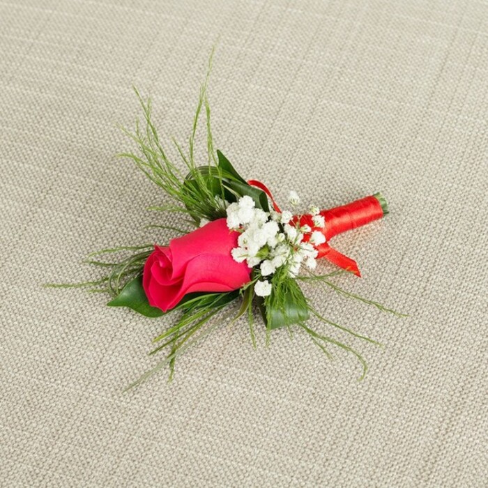 A carefully designed boutonnière rests at a slight angle on a softly textured fabric in a light beige tone, the sort of natural, understated backdrop you might find in an elegant Golders Green bridal studio. Dominating the small arrangement is a single rosebud in vivid fuchsia, leaning toward deep magenta, its petals tightly wrapped so that the bloom remains compact and full of quiet tension. The rose's surface appears silky and smooth, catching the light along the outer curves of its petals, giving a gentle sheen that emphasizes both richness and freshness. Encircling this focal flower are delicate tufts of baby's breath, each stem branching out into dozens of tiny, white blossoms that create a soft, cloud-like halo. These crisp white flecks break up the intensity of the pink and add a sense of lightness, as though the boutonnière is surrounded by a whisper of mist. Behind the rose and gypsophila, slender strands of feathery, bright green foliage stretch outward, fanning gracefully in different directions, reminiscent of the gentle, unstructured greenery you might see trailing from arrangements made for weddings in nearby Hampstead Garden Suburb. The greenery frames the rose without overwhelming it, lending the piece a natural, garden-inspired character rather than a rigidly formal look. At the base, the stems are neatly aligned and bound together with a narrow band of red satin ribbon, wound firmly and evenly to create a smooth, shining handle that echoes the richer tones buried deep within the rosebud. The ribbon does not form a large bow; instead, it is finished with a subtle, tidy fold, keeping the focus firmly on the flower itself. Clear, even lighting reveals every texture-the weave of the background fabric, the satiny petals, the airy baby's breath-imbuing the boutonnière with a feeling of freshly prepared elegance, ready to be pinned to a lapel for a wedding, prom, or refined evening event in and around Golders Green.