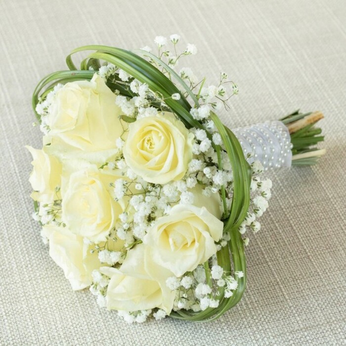 Set gracefully atop a light beige, textured fabric and basking in gentle daylight, this elegant bridesmaid bouquet is the epitome of refined floral design in Golders Green. Its composition features approximately seven to nine lush, creamy white roses, each with plush petals opening in a subtle spiral-from youthful, tightly formed buds to gracefully expanded blossoms. The roses are enveloped by generous sprays of ethereal white baby's breath, their minute blooms creating a soft, cloudlike fullness and introducing delicate contrast to the arrangement. Loops and arcs of slender, fresh green bear grass wind and weave through the bouquet, their pristine lines adding both modern structure and organic rhythm to the gathering. All stems are expertly gathered and wrapped in a sleek, dignified light grey satin ribbon, elevated by parallel rows of shimmering pearl accents-each row catching the light and offering tactile luxury. The natural stems, just visible beneath the wrap, suggest a fleeting freshness, as if clipped at Golders Green's weekly flower market. This bouquet, ideal for a bride or bridesmaid at a local ceremony near Golders Hill Park or Hampstead Garden Suburb, embodies purity, celebration, and a timeless connection to nature-its design reflecting both the traditions and tranquil spirit of NW11.