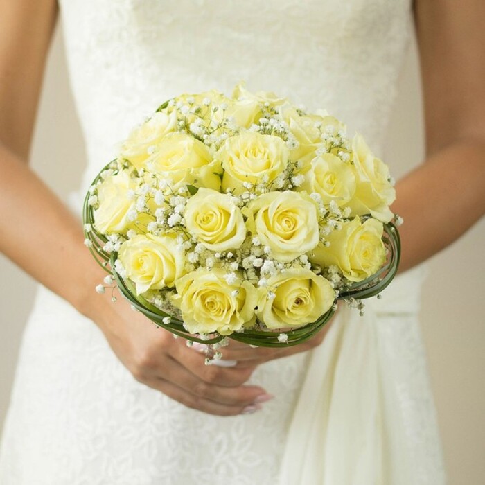 A glowing close-up reveals the delicate beauty of a bridal bouquet carried with grace by a bride dressed in an exquisitely crafted white gown, reminiscent of the classic ceremonies that take place in and around Golders Green. This meticulously arranged bouquet features lush, tightly clustered yellow roses in full bloom, their petals unfolding in soft, buttery layers that catch the ambient light drifting in from nearby Hampstead Heath. Tiny, feathery baby's breath is scattered among the roses, creating a cloud-like contrast and enhancing the bouquet's traditional appeal. Fine bands of dark green leaves or ornamental wire curl artfully around the base, highlighting the skillful design and expert touch favored by local florists. The bride's hands, polished and gentle, support the bouquet from beneath, the gleam of a wedding ring adding a subtle touch of sentimental joy. Behind her, the white fabric of a lace or embroidered bodice lends further elegance and detail, with just a hint of Golders Green ambience suggested by the gentle blur of background tones. The scene is quietly radiant, ideal for a North London love story, and offers inspiration for those seeking a combination of classic style and neighborhood character in their wedding florals.