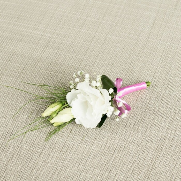 An elegant white lisianthus boutonnière is captured from above, resting diagonally across a warm, light beige fabric that resembles finely woven linen or soft burlap, its subtle cross-hatched texture giving the scene a natural, relaxed backdrop reminiscent of a refined North West London townhouse. The focal point is a single, fully opened eustoma bloom positioned slightly off-centre to the right, its many ruffled petals layered in gentle spirals, transitioning from a soft creamy base to crisp white edges. Just below and to the left, two slender lisianthus buds, still closed and tinted a pale greenish cream, lean alongside the main flower, adding a sense of progression and quiet movement, as though they might open later in the evening after the ceremony. Threaded through the composition are fine sprays of white gypsophila, the baby's breath forming tiny star-shaped clusters that create a delicate halo around the central bloom and soften the overall silhouette. From beneath the flowers, narrow, grass-like blades of deep green foliage fan out towards the lower left corner, their linear form offering contrast to the rounded petals, while one broader, darker green leaf sits discreetly behind the lisianthus, grounding the arrangement and adding depth. At the base, the neatly trimmed stems are bound together with a bright fuchsia pink satin ribbon, tied into a small bow whose smooth sheen and vivid colour stand out vividly against the muted neutrals and classic greenery. The lighting is soft and even, with no harsh shadows, allowing every petal curve, leaf vein, and thread of the woven background to be clearly defined. The result feels like a thoughtfully prepared wedding buttonhole, perhaps crafted by Flowers Golders Green for a groom or best man heading to a ceremony off Golders Green Road, combining timeless white florals with a modern pop of colour for a quietly confident finishing touch.