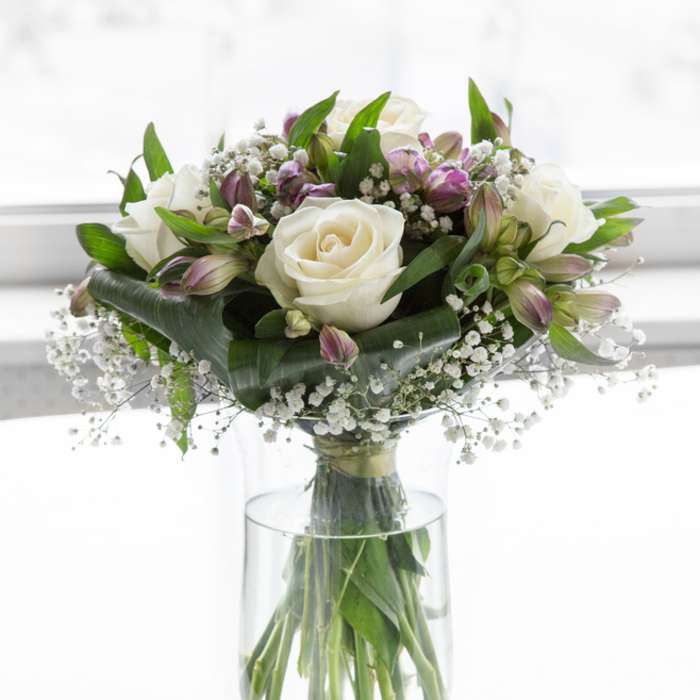 Arranged with the measured care of an experienced local florist, this tranquil bouquet stands in a simple, clear glass vase, its stems aligned in a tidy cluster that can be seen through the water. The clean lines of the cylindrical vase echo the understated mood of the flowers above. At the centre, creamy white roses open in soft spirals, each petal plush and slightly luminous in the natural light, much like petals observed on a still morning walk through nearby Golders Hill Park. Their creamy tone suggests purity and sympathy without feeling stark or cold. Threaded among the roses are elegant alstroemeria in varying stages of bloom, from green-tinged buds to blossoms tipped with violet and lavender hues. The petals of these alstroemeria are fine and gently arched, some brushed with subtle markings, introducing a tender whisper of colour that never overwhelms the arrangement's peaceful character. Tiny sprays of white baby's breath, with their airy, cloud-like texture, are woven throughout the bouquet, settling into the spaces between larger blooms and giving the whole design a delicate, almost ethereal halo. Around the base and edges, rich green foliage curves and overlaps, offering depth and contrast. Some leaves are broad and glossy; others are slender and slightly arching, creating a natural, garden-like frame. The background is very bright and softly blurred to white, as if the bouquet is set near a large window in a quiet Golders Green home, bathed in even, gentle daylight. The impression is one of calm support and respectful beauty, ideal for sending condolences or placing as a dignified focal point at a remembrance service.