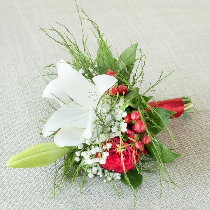 A dew-fresh, compact floral arrangement, radiantly displayed on an off-white, woven fabric surface reminiscent of boutique settings in Golders Green, blends classic floral sophistication with festive vibrancy. Commanding attention at its center is a grand white lily, petals dramatically splayed and dusted with delicate cinnamon freckles, revealing prominent golden-yellow stamens. Just below, a blooming red rose unfurls in velvet-rich spirals, bringing a touch of opulence and tradition. Baby's breath, in airy white clouds, dances between the main flowers, while glossy clusters of scarlet hypericum berries introduce a lively, celebratory pop-mirroring the spirited gatherings at local synagogues and cultural halls. Lush greens, from fine ferny fronds to broad, shadowy leaves, frame the bouquet with organic depth and textural richness. On the left, a pale green lily bud points forward, echoing the anticipation of new seasons in Golders Green's parks and gardens. Bound together with a luxe red satin ribbon, the stems complete this expertly composed piece, its balanced palette evoking romance, festivity, and heartfelt gestures. Bathed in gentle, even lighting, the bouquet's natural vibrancy is brought to life-a testament to skilled hands and local character, perfect for weddings, anniversaries, or elegant home décor delivered fresh by local florists.