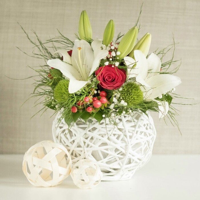 An artfully composed floral centerpiece appears as though it has just been set down on a crisp white table in a Golders Green home prepared for a wedding brunch. The arrangement is gathered in a distinctive spherical white vase, created from intertwining strands that form an open, basket-like lattice, giving it a light, contemporary feel. From this sculptural base rises a generous bouquet dominated by luxurious white lilies; some blooms are fully open, their broad petals gracefully arching back to reveal speckled throats and prominent stamens, while others remain as smooth, elongated buds, suggesting days of unfolding beauty to come. Among the lilies, a standout red rose with velvety, tightly furled petals introduces a rich, romantic focal point, supported by other deep red roses that echo its colour and intensity. Two or more bright green pom-pom flowers, likely dianthus or chrysanthemum, provide a playful contrast in both form and hue, their rounded, tufted heads adding texture and freshness. Delicate sprays of white baby's breath weave lightly through the arrangement, softening the lines and creating a cloud-like, ethereal effect. Small clusters of red berries nestle between the blooms, adding a subtle festive hint that would suit a winter wedding or holiday celebration in NW11. The bouquet is framed by abundant greenery, from fine, feathery fronds to broader, deeper green leaves that spill naturally over the edge of the vase, giving the design depth and movement. On the left side of the smooth white surface, two decorative woven balls in natural beige tones echo the vase's texture, completing the harmonious, design-conscious look. Set against a softly textured neutral background, this centerpiece feels refined and inviting, perfect for wedding table arrangements or special gatherings around Golders Green and nearby Hampstead Garden Suburb.