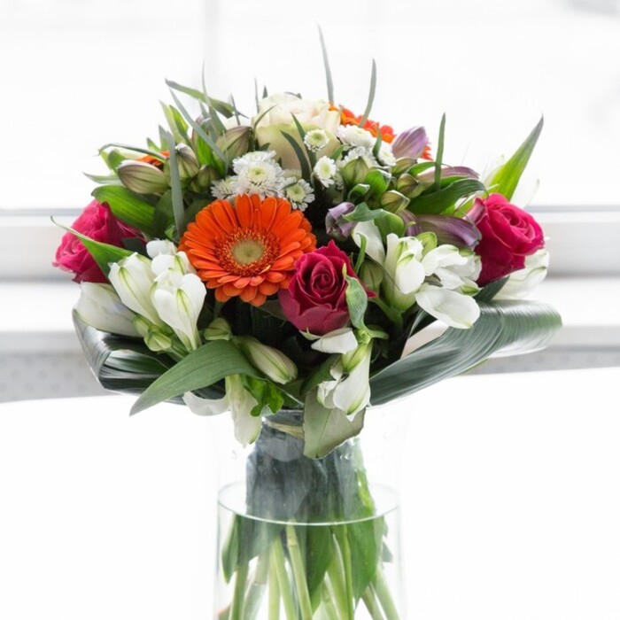 This masterful floral display, crafted by a seasoned Golders Green florist, envelops the senses with its bold blend of colour, texture, and local charm. Towering gerbera daisies in radiant orange and creamy peach hues form the heart of the arrangement, their cheerful faces highlighted by a halo of lush, emerald foliage. Rich magenta roses are woven throughout, their spiralled petals unfurling with quiet elegance, evoking the timeless romance of North End Road's secret gardens. Pristine white alstroemeria and clusters of petite spray roses nestle between the central blooms, imparting a gentle lightness that feels like the whispered promise of spring mornings at Golders Hill Park. The bouquet's structure is reinforced by broad, paddle-shaped aspidistra leaves, elegantly curled and wrapped to cradle the stems, providing both physical stability and a tranquil verdant contrast. The entire composition is presented in a slender glass vase, the fresh green stems visible beneath the water, reflecting the commitment to freshness and quality. Illuminated by diffuse daylight, the arrangement glows softly, allowing each colour and texture to sing without harshness. Suitable for any joyful gathering or to offer comfort and appreciation, this full-bodied bouquet is an authentic piece of Golders Green artistry, designed to bring warmth and optimism into any local home.