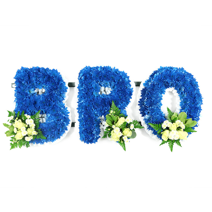 Set before a clean white background in a studio shot, this three-part floral tribute spells BRO in large, three-dimensional letters crafted entirely from saturated royal blue chrysanthemums. The blue petals are densely arranged to produce a cushiony, continuous surface with sharp, well-defined letter edges; a slim white foam frame is visible at the inner loops and along the base, indicating careful construction for standing display. Each letter is gently embellished with a small cluster of pale yellow-cream spray roses and tiny white filler blooms, positioned at the lower-left of the B and the lower-right of both the R and O. Interspersed foliage-glossy salal or lemon leaf and finer, airy fern fronds-adds contrast, texture and a soft green perfume that balances the floral sweetness. The neutral studio light renders the blue deep and velvety while letting the cream accents read as comforting and pure. This arrangement has the reverent tone suited to funerals, wakes, or memorial services, and is precisely the sort of custom tribute families in Golders Green, around Golders Hill Park and along Finchley Road might request. Prepared by Flowers Golders Green, it conveys fidelity, mourning and affection in an elegant, personal way that reads as both crafted and compassionate.