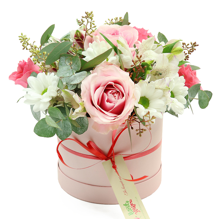An elegantly styled hatbox bouquet is displayed against a pristine white background, its colours and textures taking centre stage the way a special gift might stand out on a dining table in a Golders Green home. The container is a round, pastel pink cylinder with a matte finish, evoking both softness and sophistication. Wrapped neatly around its circumference is a narrow coral-red satin ribbon, tied into a precise, slightly glossy bow at the front. From this bow hangs a rectangular cream tag with subtle green script, hinting at a handwritten note and the personal touch of a local NW11 florist.
The flowers emerge in a full, rounded dome that gently overflows the rim of the box, creating a sense of abundance without losing its refined shape. At the heart of the composition are large, luxurious roses in pale blush pink, some with deeper fuchsia tones swirling from their centres, echoing romantic walks through nearby Hampstead Heath gardens. They are surrounded by numerous white daisy-like chrysanthemums, each with a yellow-green centre that adds a cheerful, luminous contrast to the pinks. Here and there, trumpet-shaped white alstroemeria blossoms unfurl, their petals marked with fine, speckled lines that lend a subtle, graceful pattern.
Deeper notes of colour come from smaller, vivid pink blooms-perhaps lisianthus or spray roses-tucked among the larger flowers for depth and visual rhythm. The entire bouquet is woven through with varied greenery: rounded, silvery eucalyptus leaves that suggest a fresh, aromatic scent; darker green foliage adding structure; and delicate stems bearing tiny buds or seed-like accents that introduce a natural, slightly wild charm. The result feels like a modern yet timeless gift, suitable for birthdays, anniversaries, or celebrations across Golders Green, lovingly crafted to bring a moment of joy to a home near Temple Fortune, Childs Hill, or beyond.