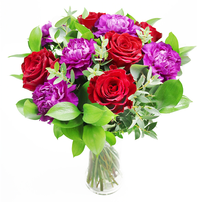 A lush, modern-classic bouquet takes centre stage in a clear glass vase, its composition carefully crafted to showcase both drama and elegance. At the forefront are velvety red roses, their petals dense and full, curling outward in perfect spirals that suggest timeless romance. Their deep crimson tones are intense but softened by the gentle glow of natural light, evoking the warmth of an evening stroll through Golders Green as shopfronts begin to light up. Interspersed with the roses are generous purple peonies, each blossom a study in texture, with layer upon layer of softly crinkled petals that range from saturated violet at the centre to slightly lighter tones at the edges. They bring a contemporary flair, like a brush of modern artistry against the more traditional roses. Encircling the blooms is a rich collar of greenery, the leaves smooth and glossy, some larger and more structured, others softer and slightly curved, creating varied rhythms of green. Slender stems of eucalyptus weave through the design, their round, silvery leaves catching points of light and adding a cool, aromatic freshness. The stems inside the vase are neatly cut and arranged with intention, forming clean, vertical lines through the clear water, giving a sense of order beneath the expressive flowers above. Light seems to fall from a nearby window, perhaps like those of a home off Finchley Road or near Golders Green station, illuminating the bouquet so the reds and purples appear almost luminous against the more muted backdrop. The entire arrangement suggests a meaningful gesture-perfect for anniversaries, romantic surprises, or to dress a dining table for a special family gathering-radiating affection, admiration, and the quiet luxury of thoughtfully chosen blooms.