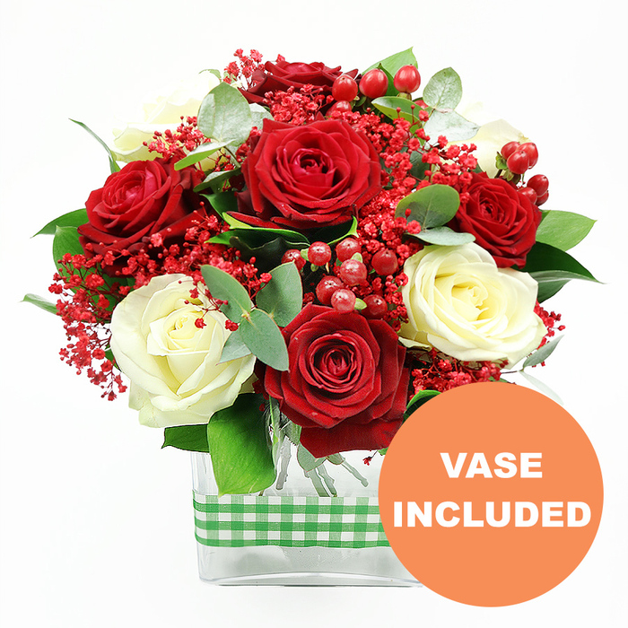 Set against a bright white background, this striking floral arrangement feels like a moment of celebration paused in time, the kind of bouquet you might place in a sunny Golders Green living room before guests arrive. At its centre, rich red roses open in plush, velvety layers, their deep colour evoking romance and evening light, while creamy white roses nestle alongside, their smooth petals forming elegant, cup-shaped blooms. The interplay between the crimson and ivory tones creates a classic, timeless contrast, reminiscent of a traditional holiday table or an intimate anniversary dinner on a quiet street off Finchley Road. Scattered generously through the bouquet are clusters of fine, bright red filler flowers, similar to tinted baby's breath, which lend a light, airy haze and soften the edges of the design. Between them, round red hypericum berries gleam like tiny ornaments, bringing a playful touch and a sense of seasonal festivity. The flowers are supported by varied greenery: broad, glossy leaves provide structure and depth, while sprigs of eucalyptus introduce cooler, silvery-green notes and a suggestion of fresh, aromatic scent. All of these elements are gathered into a clear, rectangular glass vase, the tidy stems clearly visible through the transparent sides, which gives the design a polished, ready-for-the-table appearance. Around the middle of the vase, a green and white gingham ribbon is wrapped snugly, its checked pattern adding a hint of rustic charm that would look perfectly at home in a kitchen overlooking Golders Hill Park. In the lower right corner of the image, a vivid orange circular badge with bold white text announces VASE INCLUDED, making it immediately clear that this floral gift comes complete and ready to display. The overall impression is one of thoughtful craftsmanship, warmth, and joy, ideal for same-day delivery to mark birthdays, anniversaries, or quiet gestures of love across Golders Green.