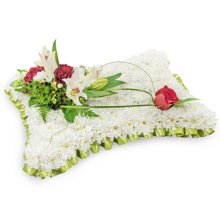 Presented against a simple white backdrop, this traditional pillow-shaped tribute reads as an expression of repose and respect. The rectangular base is completely covered with tight clusters of small white chrysanthemums, packed to create a uniformly textured surface that feels soft and quiet, like a folded linen placed at rest. A slim border of pale, lime-tinted ribbon is pleated neatly around the edge, its satiny finish catching soft light and lending the design a gentle refinement. Toward the upper left a composed spray of accent blooms rises from the chrysanthemum field: open white Oriental lilies with delicate yellow stamens and an unopened bud that suggests peaceful continuity; clusters of rich burgundy or dark red spray carnations that bring warmth and depth; and a mix of dark green foliage - including airy fern fronds and glossy leaves - that forms a verdant backdrop and adds sculptural volume. From this arrangement an elegant green arc of salal or decorative wire extends across the cushion in an open sweep, leading to a solitary deep red rosebud placed on the upper right corner, its petals just beginning to part in a quiet, dignified gesture. The overall colour story of white, soft green, and deep red conveys purity, gentle hope, and lasting affection. This arrangement would be appropriate for memorials and funeral services in Golders Hill Park area or local chapels around Hoop Lane and is crafted to offer solace through considered form, texture, and understated floral symbolism.