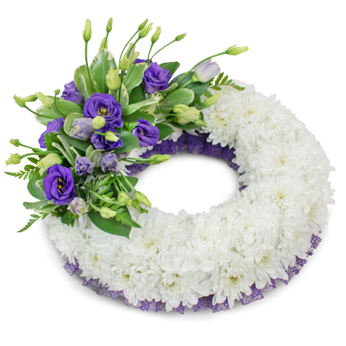 A dignified round memorial wreath composed primarily of tightly packed white chrysanthemums fills the frame, their multi-layered petals forming a plush, continuous ring that conveys purity and calm. The arrangement's visual interest is concentrated in the upper-left quadrant, where a lavish accent of purple lisianthus (Eustoma) unfolds in a range of hues from deep violet to gentle lavender; some flowers are fully open with softly ruffled faces, while others remain as poised buds, giving the cluster a natural rhythm of maturity and promise. Woven through these purple blooms is a selection of green foliage - glossy leaves with light variegation alongside delicate, feathery fern fronds - which adds structural contrast and a sense of organic movement. Along the inner and outer rims of the chrysanthemum base a narrow, crinkled strip of dark purple mesh or lace peeks out, harmonising with the lisianthus and providing a subtle, elegant border that completes the composition. The wreath rests against a clean white studio background, lit with gentle, even light that reveals the textures and casts a soft shadow underneath, producing depth without drama. This floral tribute reads as both traditional and quietly contemporary: thoughtfully balanced, tactile, and composed to communicate respect and steady remembrance. It is a suitable choice for a funeral or memorial, conveying heartfelt condolences in a manner that is graceful and composed.