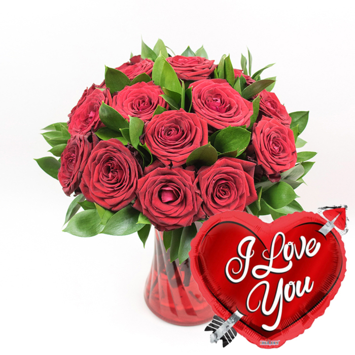 A richly romantic bouquet of deep crimson roses takes centre stage, thoughtfully arranged to convey intense affection and enduring commitment. Around a dozen or more roses form a compact, rounded cluster, their velvety petals layered generously and opening in soft spirals from the heart of each bloom. Some roses are fully open, revealing intricate petal whorls and subtle shadows that create a sense of depth, while others are partially opened, suggesting the gentle unfolding of emotion. Interlaced between the blooms, fresh dark green foliage offers a glossy contrast, its leaves catching just enough light to emphasise the richness of the red. The flowers are nestled into a clear, ruby-red glass vase, whose delicately fluted or vertically ridged sides add texture and a hint of movement as they refract light and colour. The water inside appears faintly tinted, echoing the romantic tone of the arrangement. In the lower right foreground, slightly overlapping the vase, a striking metallic red heart-shaped foil balloon adds a playful yet sincere touch. The balloon is plump and lustrous, reflecting the surroundings in soft highlights, and is boldly emblazoned with the words I Love You in white cursive lettering across its centre. A silver Cupid's arrow is illustrated as if it were passing diagonally through the heart, reinforcing the theme of love struck at its core. The entire composition rests against a crisp white background, allowing the vivid reds and fresh greens to stand out clearly and dramatically. It's easy to imagine this arrangement being delivered across Golders Green, perhaps to a flat near Golders Green station or a home overlooking nearby parks, to mark Valentine's Day, a special anniversary, or a romantic milestone that deserves a truly memorable gesture.