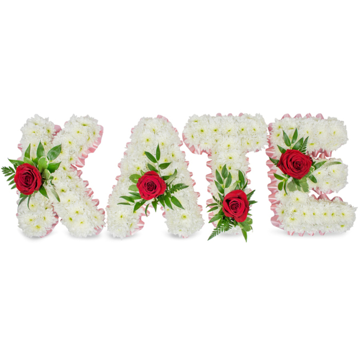 This image presents a considered and elegant floral name tribute spelling KATE in large, three-dimensional block letters, each letter painstakingly formed from a voluminous cluster of small, pristine white chrysanthemums. The chrysanthemums create a soft, pillowy texture that suggests purity and dignity, and along the outer edges each letter is finished with a subtle pale pink satin ribbon that is pleated into a neat, delicate border. A single, vivid red rose sits prominently on each letter as a bold point of contrast: on the K the rose is arranged on the lower part of its left vertical stroke; on the A the rose is positioned at the apex of the triangular peak; on the T the rose rests at the base of the left section of its horizontal crossbar; and on the E the rose graces the middle horizontal bar. Around each rose, lush green foliage - a combination of broad glossy leaves and finer fern-like sprigs - adds depth, framing the crimson blooms and softening the hard geometry of the letters.
The clean white backdrop and gentle, diffuse lighting allow the textures and colours to speak clearly: satin whisper ribbons, the chrysanthemum's layered petals, the velvety crimson of the roses and the cool sheen of the greenery. The arrangement reads as a bespoke sympathy tribute suited to funerals, memorials or remembrance ceremonies where clarity and heartfelt sentiment are required. As a local piece one might commission from a Golders Green florist working near Golders Hill Park or Hendon Lane, it balances traditional floral symbolism - white for purity, red for love - with meticulous florist craftsmanship to honour a life with quiet, assured grace.