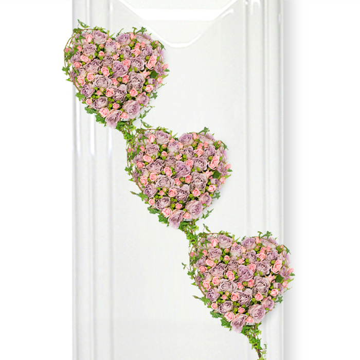 This image shows a beautifully composed sympathy arrangement of three heart-shaped wreaths, crafted entirely from roses and trailing greenery, suspended against a crisp white background that heightens both colour and form. The hearts are aligned diagonally, stepping down from the top left to the lower right, so that they appear to gently cascade, one into the next, like a sequence of shared memories. Each heart is richly packed with miniature roses in a refined palette of dusty lavender, soft mauve, and blush pink, creating a plush, velvety surface where individual blooms nestle closely together. Subtle variations in tone-from deeper purplish petals in the shadows to almost porcelain pink at the edges-add depth and a sense of quiet movement, as though the flowers were caught in soft, indirect daylight. Fresh ivy vines embrace each heart, looping elegantly around their contours and extending in arcs that link all three, giving the design a natural, almost woodland grace reminiscent of ivy-clad walls and garden corners around Golders Green and Temple Fortune. The ivy's glossy leaves provide a cool green contrast to the warm, romantic pastels, making the hearts stand out clearly while symbolising continuity and growth. Along the left of the image, translucent vertical lines hint at glass panels or a polished interior wall, suggesting a serene funeral chapel or reception area where such a tribute might be displayed. The overall effect is of a carefully considered, deeply personal piece, suited for a funeral or memorial to express enduring love between generations, or to honour a cherished partner, parent, or close friend from the Golders Green community.