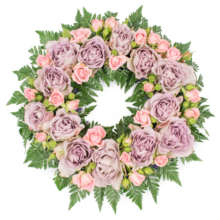 Laid flat against a crisp white background, this floral wreath forms a flawless circle, its carefully balanced proportions speaking of a skilled florist's hand. The entire ring is thick with roses, creating a cushion of petals that feels both luxurious and deeply gentle, the sort of tribute families in Golders Green often choose to say a final farewell. Dominating the design are generously sized roses in dusty lavender and soft mauve, fully open so that every layered petal is visible, from the gently cupped outer edges to the softly shadowed centres. Their colour is nuanced-a greyish-purple that feels antique and timeless, perfectly suited to a moment of reflection. Threaded among these larger blossoms are clusters of smaller blush-pink spray roses, some in tight bud, others at various stages of opening. This mix of closed and half-open blooms adds movement and texture, suggesting memories unfolding and the tenderness of shared moments. Beneath and around the roses, a dense halo of dark green fern fronds forms a strong, verdant base. The ferns' fine, toothy leaves fan outwards, creating a textured border that frames the pastel tones and gives the wreath a grounded, natural look, as though it could have been gathered from a shaded garden just off Golders Green Road. Here and there, you can glimpse deeper filler greens and tiny darker buds tucked between the flowers, adding depth and a sense of quiet complexity. The lighting is even and calm, highlighting the interplay of purple, pink, and green without cast shadows. The overall impression is one of calm reverence and heartfelt sympathy-a classic funeral or memorial wreath symbolising continuity, remembrance, and the comfort of lasting love.