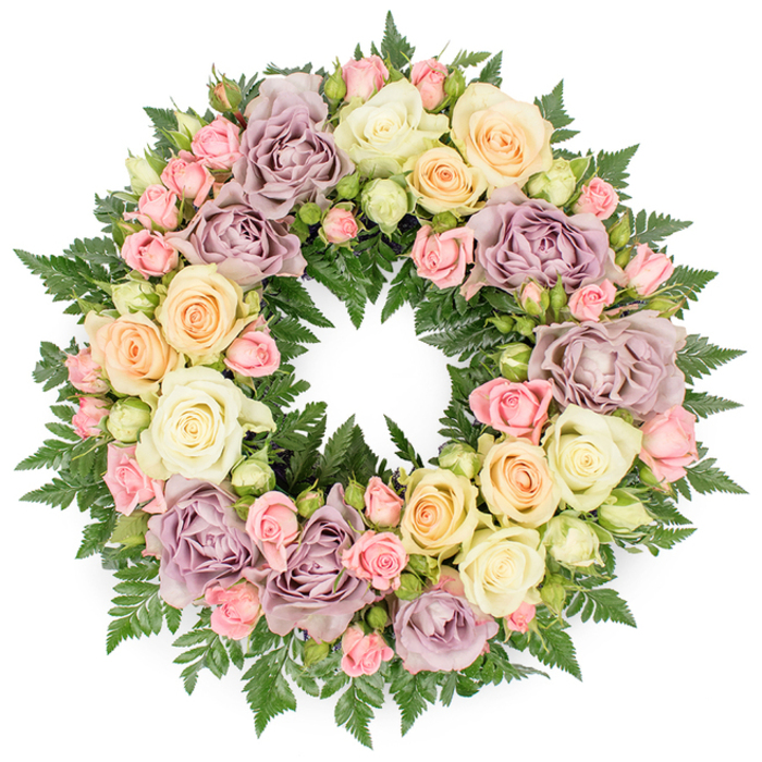 A classic funeral wreath, photographed head-on against a bright, white backdrop, shows a complete circle of roses and ferns so densely arranged that the underlying frame is invisible. The composition is rich but not overpowering, built around generous, fully open roses in soft pastel tones: creamy white, light peach, and warm ivory are accented by unusual dusty lavender and muted mauve blooms, giving the tribute a refined, almost old-English garden character. Threaded throughout are spray roses in tender blush pink, many of them still in bud or just beginning to open, alongside pale green rosebuds that add a sense of gentle movement and growth. Fine, dark green fern foliage forms the structural base and peeks out from between the petals at regular intervals, adding cool shadows, texture, and a deep contrast that makes the paler roses glow. The circle is unbroken and symmetrical, symbolising unending love and remembrance, the kind of design that might be chosen for a service in Golders Green after a quiet family gathering on nearby Finchley Road or around Golders Hill Park. Soft, even lighting allows the viewer to appreciate the satin-like petals, the ruffled edges of the blooms, and the feathery fronds of the ferns. The overall impression is of a thoughtfully crafted tribute, combining serenity, respect, and a comforting sense of continuity, ready to be placed beside a coffin or at a memorial as a final, elegant gesture of farewell.