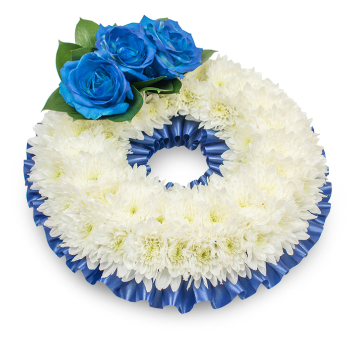 Viewed against a pristine white backdrop, this handcrafted circular wreath presents a composed interplay of texture and colour designed for moments of solemn remembrance around Golders Green and nearby Finchley Road. The main body is a full, domed circle of immaculate white chrysanthemums, their multitude of delicate, feathery petals building a soft, tactile mass that reads as serene and pure from any angle. On the upper-left portion three striking deep-blue roses form the arrangement's heart: each rose holds densely layered, velvety petals in an intense royal to cobalt hue, their richness amplified by the surrounding glossy dark-green foliage. A pleated satin ribbon, dyed to match the blue of the roses, borders both the outer and inner edges, its ruffled folds catching the light and adding a refined, ceremonial frame to the floral volume. The clean composition casts a subtle shadow on the white surface beneath, giving the wreath a gentle three-dimensional presence as though ready to be carried or positioned near a memorial display. The sensory impression is quietly tactile - the cool silkiness of the ribbon, the soft papery feel of chrysanthemum petals, and the hushed, floral whisper of the blooms - all chosen to convey dignity, solace, and a personal note of respect. I imagine this piece laid with care at a chapel entry near Golders Hill Park or placed beside a memorial tablet, where its calm palette and craftsmanship offer consolation to mourners.