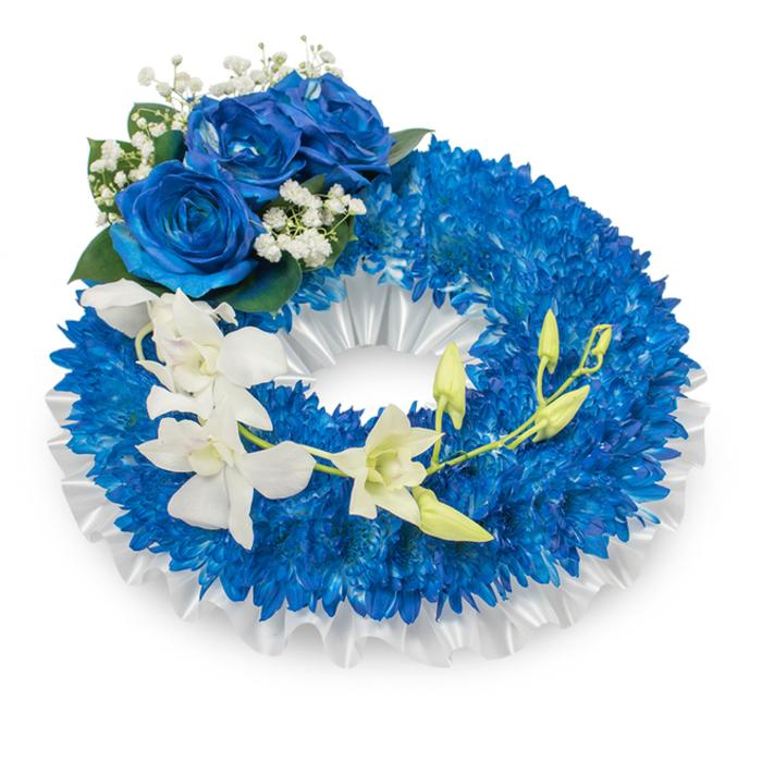 An elegant sympathy wreath combining saturated blues and pristine whites, described with the detail of a florist who crafted it in a small Golders Green studio. The circular form is densely faced with tiny royal-blue chrysanthemum florets; each little bloom contributes to a continuous, tactile ring that feels full and measured, the petals catching a cool, diffuse light to emphasize depth. Underneath and gently visible at the inner edge lies a ruffled white satin ribbon that has been pleated and secured to form a soft, luminous frame-the ribbon's smooth sheen offsets the chrysanthemums' fine texture and lends formal structure. At the top-left curve, three substantial blue roses are arranged as a refined cluster: their petals unfurl in deep sapphire layers with a plush, velour-like surface, accented by delicate sprays of white baby's breath and touches of deep green foliage for balance. From mid-left, a slender stem of ivory orchids drapes gracefully toward the lower-right, the broad, silky petals and subtle throat markings offering an airy counterpoint; several lime-green buds sit among the open flowers, poised to bloom and symbolise renewal. The composition is finished with discreet wiring and stem-support to preserve its sweeping line, and the overall impression is one of quiet respect, calm, and meticulous craftsmanship. Suitable for memorial services around Golders Green Road, near Golders Hill Park or Hampstead, this wreath conveys sympathy through colour, texture and considered floral technique.