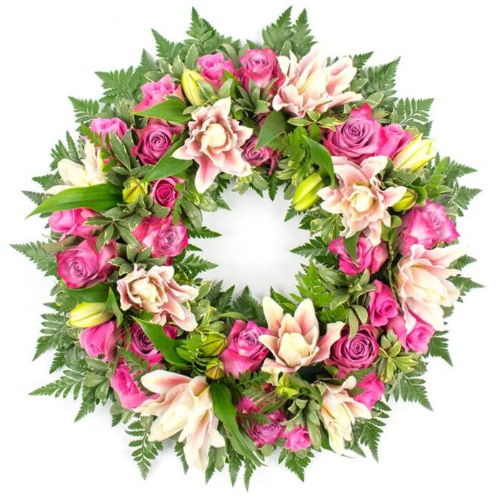 Set against a spotless white backdrop, this circular funeral wreath appears almost to float, a complete ring of flowers and foliage symbolising an endless bond. The wreath is generously packed with roses in layered tones of intense pink: deep magenta blooms with velvety petals, fuchsia roses that catch the light, and softer blush buds just beginning to unfurl. Between these romantic roses sit full, double-petalled lilies in creamy white and delicate pastel pink, some edged with a whisper of darker pink along their tips and veins. A few younger lily buds in vivid lime-green push upwards, their pointed forms suggesting new life amid remembrance. The flowers rise from a deeply textured collar of greenery, including dark, feathery fern fronds that create a soft fringe around the outer edge and polished leaves that vary from broad and oval to narrower, more architectural shapes. Subtle variations in leaf colour-from dark forest green to brighter, fresher hues-add natural depth and structure, preventing the wreath from feeling flat. The evenly diffused lighting highlights the intricate ruffles of each petal and the gentle gloss on the leaves, without harsh shadows. Visually, the design feels calm, carefully balanced, and thoughtfully composed, just the kind of tribute you might see delivered to a service in Golders Green, near the crematorium or a local synagogue, where families gather to share memories. This wreath offers a refined yet comforting expression of sympathy, suitable for funerals, memorial gatherings, or to rest by a grave as an enduring sign of respect, love, and gratitude.