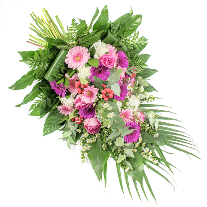Set against a crisp white background, this substantial funeral spray is arranged on a gentle diagonal, beginning with a compact tie of natural stems at the upper left and sweeping downwards into a trailing arc of leaves and blooms at the lower right. The colour harmony centres on pinks, magentas, and whites, with several large gerbera daisies in pale blush and vivid fuchsia forming the main focal points; their round faces and finely layered petals give the tribute a sense of openness and light. Between them nestle classic pink roses in various stages of opening, some still tightly budded, others just beginning to reveal deeper inner petals, suggesting a life remembered through many chapters. Soft white flowers, similar to miniature carnations or dianthus, cushion the bolder tones with their ruffled edges, while tiny white daisy-like blossoms are scattered throughout, lending a wildflower charm reminiscent of the verges near local streets like Golders Green Road and Hoop Lane. Here and there, clusters of red hypericum berries emerge from the greenery, their smooth, bead-like forms adding texture and a rich, berry-red note. The arrangement rests on a luxurious bed of foliage: broad, glossy dark green leaves create a strong base; bright green, feathery ferns soften the silhouette; and sprigs of rounded, silvery eucalyptus thread through the flowers, implying a cool, gently aromatic fragrance. Long, slim palm leaves extend from the lower right, accentuating the cascading motion and giving the spray a finished, polished outline. Crafted with the care of an experienced Golders Green florist, this design works beautifully as a casket spray or standing tribute at a memorial, offering a dignified, heartfelt expression of condolence and a serene celebration of a cherished life.