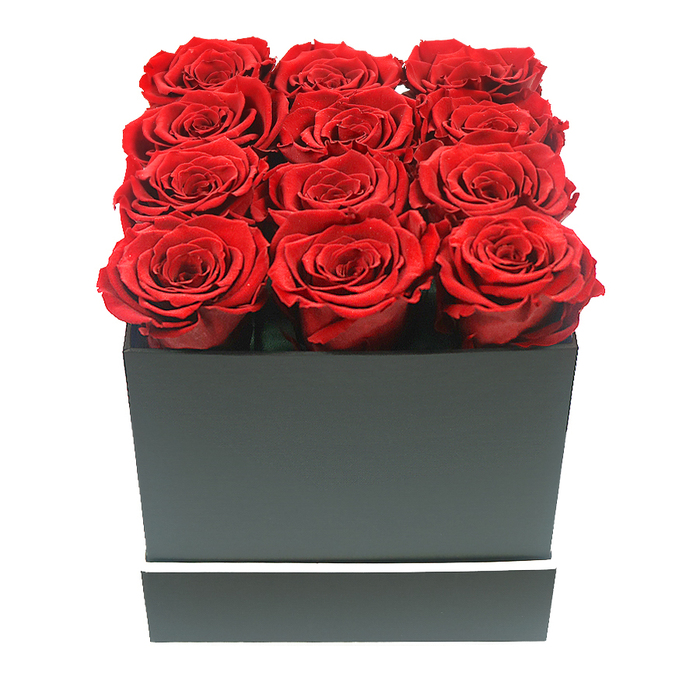 A refined, studio-style photograph showcases a premium arrangement of twelve preserved red roses presented in a modern square gift box, captured in clear detail as if placed on a white tabletop in a stylish Golders Green apartment. The roses form a perfect 3x4 grid, tightly packed so that no gaps are visible between the blooms. Each flower is in full, rounded bloom, with layers upon layers of velvety petals curling outwards from a deeper heart of crimson, giving a plush, cushion-like appearance. Their hue is a rich, commanding red, with subtle tonal shifts that create a sense of depth and movement, almost like ripples of silk. The blooms rise slightly above the edges of a sleek, matte black box, whose clean lines and square shape suggest contemporary floral design often requested for chic celebrations and romantic surprises around Golders Green and Temple Fortune. The surface of the box is smooth and untextured, absorbing light softly, while a slender white stripe encircles the lower part like a minimal decorative band, offering a crisp contrast that lifts the overall presentation. The background is an unbroken field of pure white, and soft, even lighting gently illuminates the roses from above, creating delicate shadows between the petals and along the box's sides. This subtle play of light emphasises the sculptural quality of the arrangement without any distraction. The composition radiates quiet opulence and enduring affection, the kind of gift one might send through local same-day delivery to surprise a loved one near Golders Hill Park or a friend hosting a dinner on Dunstan Road. The photograph speaks of lasting romance, carefully considered gifting, and the timeless allure of red roses that never fade.