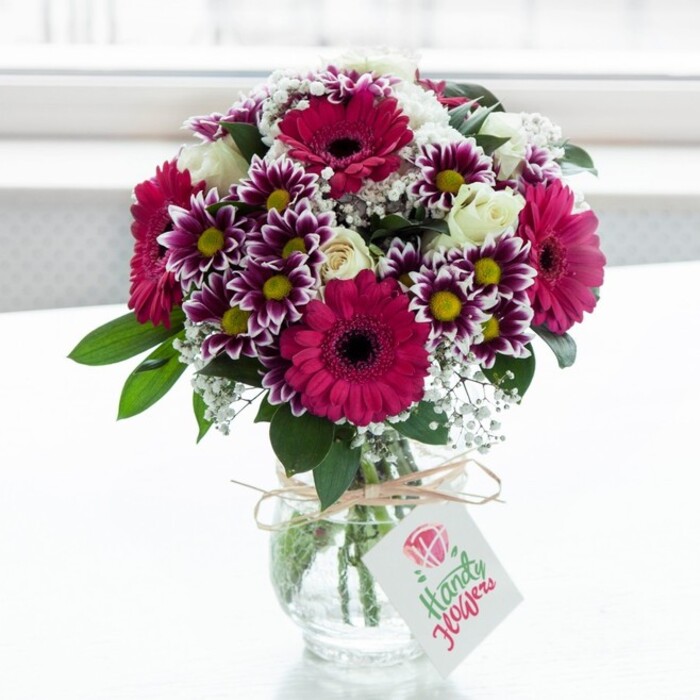 A lush, exuberant bouquet of mixed flowers is displayed in a clear, jar-shaped glass vase, set against a luminous white backdrop that recalls soft daylight pouring through a Golders Green bay window. The arrangement is dominated by large gerbera daisies in intense shades of magenta and deep pink, each bloom with a dark, almost chocolate-coloured centre that adds depth and drama. Surrounding them, intricate bicolour chrysanthemums offer a striking contrast: their many petals are tipped in rich purple that gradually dissolves into pure white, encircling bright yellow centres that catch the light. Interspersed throughout are creamy white roses and delicate spray roses, some in full bloom and others still in bud, introducing an elegant, timeless note that softens the vivid palette. Fine clouds of white baby's breath (gypsophila) lace between the larger flowers, creating a light, airy texture that lifts the entire composition, while glossy dark green leaves fill out the base and edges, giving the bouquet a sense of fullness and natural movement. The stems are cleanly cut and arranged with care, visible through the transparent vase, whose neck is wrapped in a simple coil of natural twine tied into a small bow. A pale rectangular gift tag hangs from the twine, decorated with a stylised pink rose icon and flowing green script, suggesting a personal message attached for the recipient. The soft, even lighting enhances every petal and leaf, making the bouquet appear freshly arranged, as though it has just been delivered from a local florist near Golders Hill Park or the high street in Golders Green, ready to brighten a birthday table, anniversary dinner, or thoughtful thank-you gesture. The overall mood is joyful yet refined, a carefully crafted floral gift that brings colour, texture, and warmth into a London home.