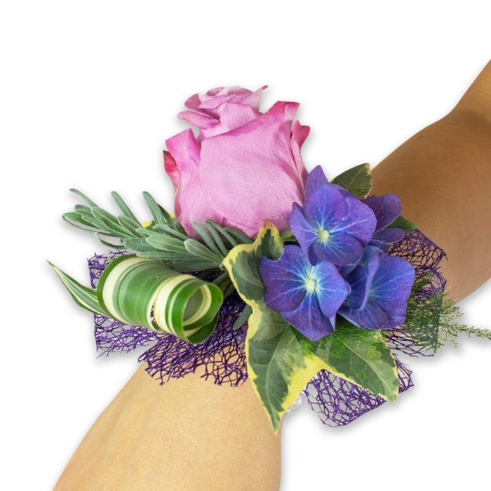 An elegant wrist corsage sits gracefully on a fair forearm, photographed on a stark white field that highlights every hue and contour. The dominant bloom is a generous, partially open pink rose-its petals moving from a richer fuchsia at the ruffled outer edges to a softer, tender pink inward-creating a romantic, vintage-inspired focal point. Flanking the rose are three dense clusters of deep indigo-purple hydrangea; their many small petals gather around pale, almost glowing centres, producing a textured, luminous contrast that reads like dusk captured in flower form. The greens are varied and intentional: long, slender silvery-green sprigs, akin to eucalyptus or lavender, form a soft, whispering backdrop; glossy variegated ivy leaves, edged in cream and pale yellow, bring a classic botanical frame; and a distinctive broad, striped leaf is curled into an artful spiral, its bold white-and-green bands lending a contemporary sculptural note. Underneath, a dark purple open-weave mesh peeks through, providing depth, subtle sheen, and cohesion with the hydrangea's tones. The arrangement balances softness and structure-silky rose petals, velvety hydrangea heads, crisp ivy and airy mesh-making it a sophisticated choice for proms, weddings or special receptions. Crafted with attentive hands and an eye for lasting beauty, this corsage evokes the gentle excitement of an evening out in Golders Green or a nearby celebration in Hampstead Garden Suburb, where handcrafted touches and fresh local floristry matter.