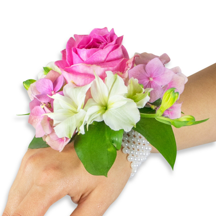 An expertly arranged wrist corsage lies against a pristine white background, its craftsmanship evident in every layered petal and leaf. At the core is a generous, fully opened hot fuchsia-pink rose - petals dense and velvety, curving outward in a classic spiral that draws the eye. Soft, pastel-pink hydrangea florets surround the rose, building a plush, textured foundation whose delicate surfaces catch light with a gentle glow. Interspersed among the hydrangea are crisp white alstroemeria (Peruvian lilies), each with four to six petals and subtle light-green streaking at their centres that lends a fresh, botanical clarity. A small cluster of pale green alstroemeria buds and a few barely unfurled pink buds sit to the right, suggesting movement and promise, while broad, rich emerald leaves frame the arrangement and provide contrast. The entire composition is secured to the left wrist of a wearer with a light-to-medium skin tone by a refined, multi-strand bracelet of tiny white pearls; the pearls form a delicate band that wraps comfortably around the wrist and extends slightly over the back of the hand, their soft lustre complementing the blossoms. The light is even and soft, accentuating textures rather than harsh shadows, and the overall mood is elegant and romantic - suitable for weddings, graduations, or prom evenings. Prepared with neighbourhood care by Flowers Golders Green, this corsage embodies the attentive floristry and local sensibility you'd expect from a Golders Green florist crafting pieces for celebrations near Golders Hill Park and Golders Green Road.