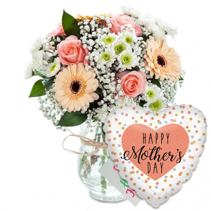 This image portrays a joyful Mother's Day gift ensemble arranged on a pristine white backdrop, reminiscent of a bright window display along Golders Green Road. On the left stands a full, rounded bouquet of fresh flowers, gathered in a clear glass jar-style vase whose neck is adorned with a casual tie of pale brown twine, suggesting a thoughtful, hand-finished touch. The bouquet's colour palette is soft yet vibrant: several peach and light pink roses are captured in mid-bloom, their petals gently unfurling with a velvety texture, while two prominent peach gerbera daisies with dark brown centres add a bolder, graphic element. Weaving between these focal flowers are light, cloud-like tufts of white baby's breath, giving the arrangement an airy, almost floating quality. Small, white daisy-shaped chrysanthemums, each with a pale green centre, nestle into the spaces, increasing the sense of lush fullness, like a garden bed in full spring flourish behind the houses near Golders Green station. Lush green foliage surrounds and supports the blooms, creating depth and framing the pastel petals with rich natural tones. To the lower right of the vase, overlapping its base, a large heart-shaped foil balloon leans into the composition. It shines in clean white, scattered with bright gold and silver polka dots that catch the light as if twinkling under shop lamps. At the balloon's centre sits a solid peach heart, inside which the words HAPPY Mother's DAY are printed in crisp black lettering: HAPPY in a simple, bold font, and Mother's DAY in an elegant, flowing script that adds a personal, handwritten feel. A small white greeting card peeks from behind the balloon, reinforcing the sense of a complete Mother's Day gift. The overall effect is one of warmth, celebration, and gratitude, ideal for illustrating Mother's Day bouquets available for local delivery throughout Golders Green.