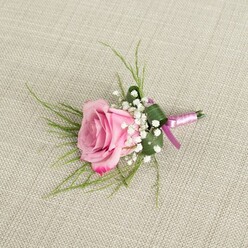 An elegant pink rose boutonniere is displayed on a neutral, light beige linen fabric, the woven texture of the cloth visible beneath the flowers like the calm backdrop of a dressing room near Golders Green station just before guests head out to a ceremony. At the centre, a single semi-open blush rose glows with soft romance, its velvety petals unfolding in gentle layers from a pale, almost porcelain pink at the core to a more saturated rose-pink at the curled, slightly ruffled outer edges. The bloom feels freshly cut, as though it has just arrived from an early-morning market delivery, with a quiet dewy sheen that hints at subtle fragrance. Encircling the base of the rose, delicate sprays of white baby's breath cluster into airy tufts of tiny blossoms, each one like a miniature pearl, lending a cloud-light contrast and enhancing the boutonniere's classic, timeless character. Slender, grass-like blades of bright green foliage arc outward from behind the flower head, adding a sense of lightness and flow, reminiscent of the gentle movement of leaves along nearby Hampstead Heath in a breeze. Nestled beneath, a broader, darker green leaf is folded with almost origami-like precision around the lower half of the stems, creating a neat, architectural cradle that keeps the design compact and refined. The stems themselves are wrapped in a smooth satin ribbon, combining a soft lavender-pink with a deeper magenta-purple edge, the colours blending harmoniously with the rose and tied in a modest knot that lies flat and secure. The natural light in the image is gentle and even, casting faint, diffuse shadows that give depth to each petal and sprig. The overall mood is one of quiet celebration and poised anticipation, a small yet meaningful floral accent perfect for weddings, proms, or formal events across Golders Green and the surrounding North West London neighbourhoods.