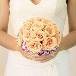 This close-up wedding image captures the midsection of a bride, her classic ivory gown forming a soft, luminous canvas for the bouquet held just above her waist. Delicate lace embellishes the fitted bodice, tracing a graceful V-neckline, while the skirt falls in smooth, gently rippling layers, suggesting movement even in stillness. Nestled between her lightly clasped hands is a meticulously arranged, round bouquet that commands the viewer's attention. At its centre, a multitude of roses in soft peach, apricot, and blush tones are packed closely together, their petals unfurling in plush, velvety spirals. The colours are warm and tender, reminiscent of a late summer sunset over nearby Golders Hill Park. Beneath this dome of roses, a striking ring of ruffled flowers in deep, royal purple fading into creamy white edges, likely lisianthus, frames the arrangement like a delicate frill. These cooler tones create a refined contrast, making the warm roses appear even more radiant. Threads of airy white baby's breath weave in and out at the base, their tiny blossoms giving a light, cloudlike texture. The bride's nails are neatly manicured in pale, neutral shades, with a subtle glitter on one thumb adding a personal, modern twist to her otherwise traditional look. The overall mood is one of poised anticipation-a bouquet carefully chosen from a local Golders Green florist, ready to be carried through synagogue doors on Dunstan Road or into a reception just off Golders Green Road. It is a design that balances romance and individuality, perfect for wedding photographs that will be cherished for years.