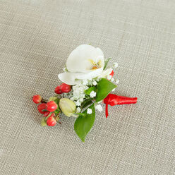 A refined boutonniere rests gently on a pale oatmeal-coloured, woven fabric background that resembles finely textured linen, its neutral tones reminiscent of the understated elegance often seen at classic Golders Green celebrations. At the centre of the design, a flawless white Phalaenopsis orchid opens gracefully, its smooth, sculpted petals creating a serene focal point. Within the orchid's throat, a subtle blend of creamy yellow and soft orange adds warmth, like a tiny flame glowing at the flower's heart. Encircling this central bloom, delicate sprays of white baby's breath form a soft, cloud-like ring, their tiny, starry blossoms adding both volume and an ethereal, romantic air. On one side, a lively cluster of glossy red berries introduces a vivid splash of colour, their rounded shapes catching the light and suggesting festivity, while a few lighter green buds nestled nearby hint at new beginnings. Interspersed through the arrangement are fresh green leaves, some broad and pointed, others slim and tapering, creating layers of texture and framing the orchid with natural grace. The stems are gathered and meticulously wrapped in a rich red satin ribbon, the fabric smooth and slightly lustrous, tied into a small, secure knot with short, elegant tails that sit flat against the background. The lighting feels like gentle daylight, revealing every vein in the foliage and the satin sheen of the ribbon without harsh shadow. This floral piece carries the quiet promise of an important moment - ideal for a groom walking out of a house near Golders Hill Park, a prom date meeting on Finchley Road, or any formal occasion in Golders Green where a single, beautifully made buttonhole can speak of care, style, and heartfelt intention.