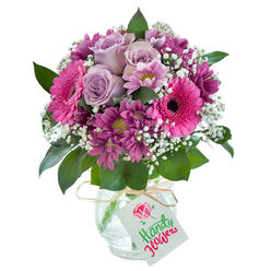 Radiating heartfelt warmth and local charm, this densely layered bouquet bursts with colour and emotion in the heart of Golders Green. At its centre, several lavender-hued roses unfurl their petals slowly, as if caressed by gentle sunlight streaming through a window on Princes Park Avenue. Encircling these roses, bold fuchsia Gerbera daisies make a statement-each blossom wide open, their dark cores like a cheerful wink to passersby browsing Golders Green High Street's abundant markets. Velvety, magenta chrysanthemums nestle in between, each flower a soft cluster echoing the deep, inviting shadows found beneath the old beech trees of Childs Hill Park. Bright flecks of white baby's breath bring lightness and texture, their tiny blossoms weaving a cloud-like effect that softens the whole ensemble. Rich, dark greenery tumbles around the florals, their glossy leaves reminiscent of a fresh morning stroll through Hampstead Garden Suburb. The arrangement sits elegantly in a textured glass vase, catching sunlight and casting fractured reflections like the pond at Golders Hill Park. A natural raffia tie encircles the vase's neck, holding a blank white tag-ready to carry a birthday wish, a note of thanks, or simply 'I'm thinking of you' from your neighbours in NW11. Each element evokes a gentle yet celebratory mood, a tangible expression of thoughtful gifting shaped by the skilled hands of Golders Green's local florists.