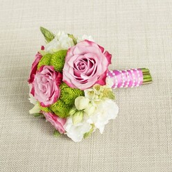 Resting on a light tan, woven fabric that looks much like a linen tablecloth you might find in a cosy Golders Green café, this compact bouquet is arranged with the care of a bespoke bridal piece. The eye is immediately drawn to three lush pink roses, their petals unfurling in generous spirals. Each bloom carries a soft, powdery blush at its heart that gradually deepens into a more intense magenta along the tips, creating a gentle shaded effect that feels both romantic and refined. Around these roses, clusters of vibrant lime-green button chrysanthemums sit like tiny cushions, their rounded forms and densely packed florets giving the posy a playful, tactile dimension. Between the green pom-poms, delicate white and cream blossoms-suggestive of lisianthus, freesia, and fine star-shaped wax flowers-thread through the design, lending a sense of lightness and a whisper of fragrance. Small touches of greenery anchor the palette and echo the fresh, garden-like charm of local parks. The stems are neatly aligned and hand-tied, then wrapped in a vivid fuchsia satin ribbon that gleams softly. Over this, a narrow strand of petite white faux pearls has been wound in an elegant diagonal pattern, as if a bracelet had been lovingly draped around the handle. The trimmed green stems extend just past the ribbon, tidy and deliberate. Soft, even lighting reveals every texture without harsh shadows, highlighting the bouquet's balance of colour and form. It feels perfectly suited to a bridesmaid's walk through a synagogue wedding in Golders Green, an intimate civil ceremony, or a stylish engagement dinner at home, offering a blend of classic romance and contemporary London flair.