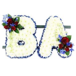 A carefully composed funeral tribute spells the letters B and A in bold uppercase, each letter formed from hundreds of compact, pristine white chrysanthemum florets to create a soft, textured surface that reads as serene and respectful. The edges are framed with a neat, ruffled royal blue satin ribbon, which crisply defines the shapes and introduces a formal, almost heraldic contrast to the white mass. On the upper left curve of the B and the lower right flank of the A sit matching accent clusters: rich, deep red roses in several stages of bloom paired with vivid blue, delphinium-like flowers that offer cool height and a contrasting pop of colour. Dark green foliage and pale green trailing stems are woven through these groupings, adding gentle movement and a natural, sympathetic softness. The two letters rest side-by-side on a slim dark green horizontal base, a freestanding presentation intended for chapel, church hall or remembrance service display. Under soft directional light the chrysanthemums glow with a calm radiance, the roses read as warm declarations of love, and the blue accents provide a tone of dignity. Crafted with care in the Golders Green neighbourhood, this style of bespoke letter tribute is what families request when they want initials to feel both personal and formal. Flowers Golders Green prepares and can deliver similar memorial pieces across the local area, including deliveries toward Finchley Road and nearby Hampstead Garden Suburb, designed to convey honour, memory and a considered farewell.