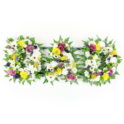The image presents a heartfelt DAD funeral tribute, crafted entirely from fresh flowers and foliage and set against a bright, uncluttered white background that makes every petal stand out. Three robust, freestanding letters - D, A, and D - stretch in a neat row, each one framed in a generous collar of lush green fern, the fronds gently ruffled as if stirred by a passing breeze on Finchley Road. Inside this verdant edging, the florist has layered a dense carpet of white blooms: open-faced white gerbera daisies and rounded chrysanthemums press closely together, their soft textures creating a cushion-like effect that feels both comforting and serene. Between the whites, cheerful yellow gerberas and petite yellow spray roses are tucked in, adding small bursts of sunlight that hint at happy memories and shared laughter. Pops of purple lisianthus, rich fuchsia spray roses, and clusters of lavender-toned flowers introduce more depth, echoing the bittersweet mix of sorrow and gratitude that often accompanies a farewell. Small, bright green button chrysanthemums add a fresh, modern touch, catching the eye without disturbing the balance of the design. Discreet metal or dark green connecting bars hold the letters in a perfectly straight line, so the tribute reads clearly from a distance at a funeral service, memorial, or wake in and around Golders Green. The arrangement has a timeless, traditional feel, yet it also reflects the care and sensitivity of a local florist used to guiding families through loss with gentle, personal touches.