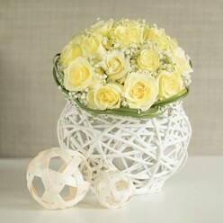 A gentle, sophisticated floral scene unfolds on a pristine white tabletop, where a rounded bouquet of pale yellow roses sits poised as if awaiting guests at an elegant Golders Green reception. The roses are tightly massed into a perfect dome, their petals curled and layered in soft buttery tones that suggest early morning sunlight filtering through curtains. Between the blooms, dainty sprigs of white baby's breath weave in and out, like fine lace threaded through the design, adding both airiness and a delicate texture that contrasts with the plump, velvety rose heads. A narrow belt of glossy green foliage hugs the base, giving a sense of structure and subtly framing the flower heads, much like a ribbon cinching a perfectly tailored dress. The arrangement is cradled in a sculptural white woven vase, its openwork pattern made of slender, interlaced strands that resemble contemporary wicker art. Light slips through the gaps, projecting faint, intricate shadows onto the white surface below. To one side sit two decorative woven orbs in pale natural hues, their looping lines mirroring the vase and tying the composition together. Behind it all, a soft, neutral background gently fades away, ensuring the viewer's attention lingers on the interplay of cream, white, and green. The image feels calm and curated, ideal for envisioning a wedding table in a quiet venue near Golders Green station or a stylish family celebration close to Hoop Lane, where flowers are chosen not just for colour but for mood and meaning.