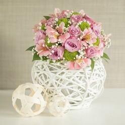 A sculpted floral centerpiece sits confidently on a spotless white surface, the kind of arrangement a local Golders Green florist might place in a calm, light-filled sitting room off Finchley Road. At its heart is a distinctive white vase made from slender, intertwined strips that form an open, basket-like lattice. The rounded body and slightly narrower neck give it substance and stability, while the airy design lets soft shadows flicker across the table. From this textured base rises a generous, rounded bouquet that feels both luxurious and approachable. Roses in pastel pink and gentle lavender are fully open, their velvety petals layered in delicate whorls that invite the fingertips as much as the eyes. Interwoven among them are stems of pink alstroemeria, each bloom with trumpet-shaped petals etched with fine stripes and subtle markings, hinting at yellow or deeper pink in the centre. Lime green button chrysanthemums punctuate the softer shades, each small, spherical flower head composed of tight, neat petals that bring a crisp, contemporary pop of colour. Wisps of white baby's breath thread through the bouquet, filling small gaps and casting an airy, cloud-like veil that softens the transitions between blooms. Dark green foliage hides in the depths of the arrangement and peeks out at the edges, providing contrast and natural structure. To the left of the vase, two decorative spheres made from pale, woven strips are placed with care, one slightly larger and set back, the smaller one nudged forward, echoing the vase's openwork and adding a crafted, homely touch. A softly textured beige backdrop and gentle, even lighting complete the scene, creating an atmosphere of tranquil celebration that would suit a quiet anniversary dinner or thoughtful thank-you gift delivered from Flowers Golders Green across NW11.